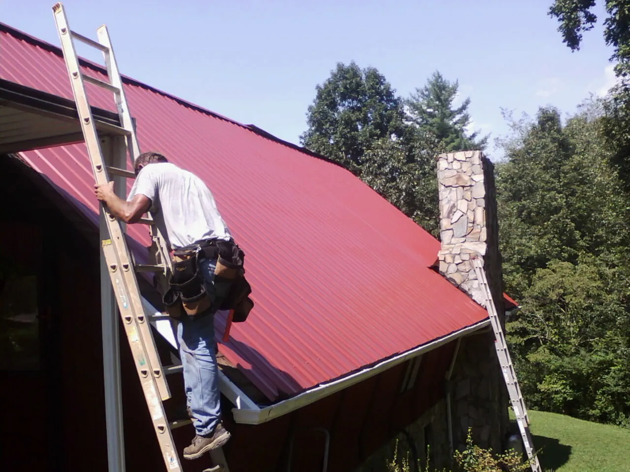 Roofer on a ladder beside a red metal roof and stone chimney — Soffit & Fascia Repair by Roofing Brothers in Biddeford Pool